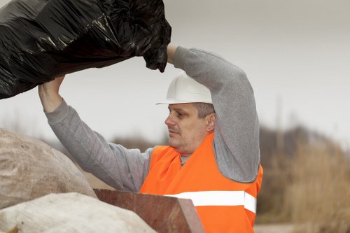 A male waste removal worker wearing a white safety helmet and an orange high-visibility vest is standing outdoors in a rural or semi-rural setting, with blurred natural background elements. He is holding a large, black plastic rubbish bag in both hands, lifting it above stacked beige and brown sacks or waste containers. The worker's expression appears focused on the task. The scene suggests a rubbish collection or waste management activity, consistent with services provided by Commercial Waste Removal Chingford, in an environment typical of a rural or suburban area near Chingford, with natural outdoor lighting and an overcast sky.